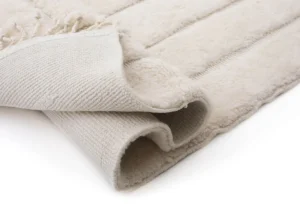 A folded beige luxury rug with a textured underside, featuring layered seams and fringed edges.