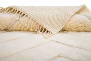 A close-up of a cream-colored throw with a textured surface and tassels along the edge, elegantly draped over another soft, fuzzy blanket.