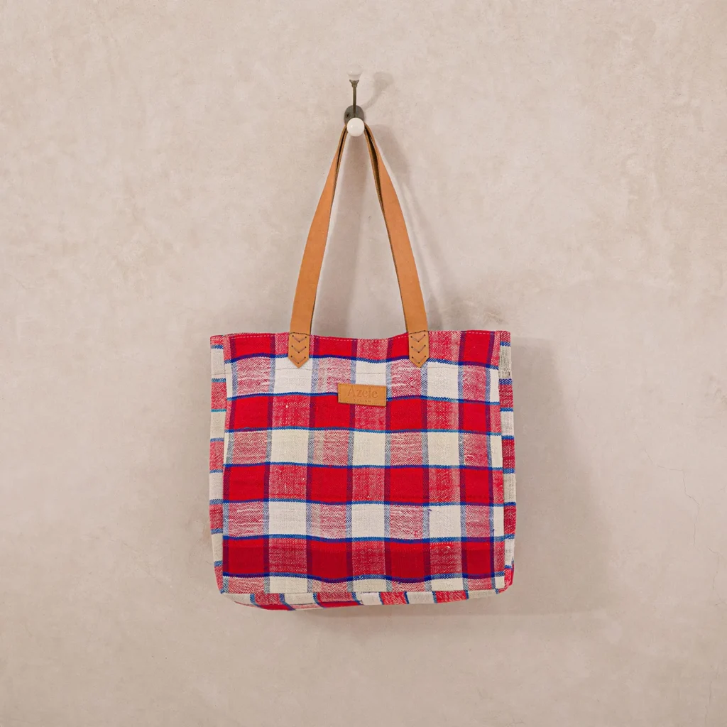 A red and white plaid tote bag with brown leather handles hangs on a wall hook.