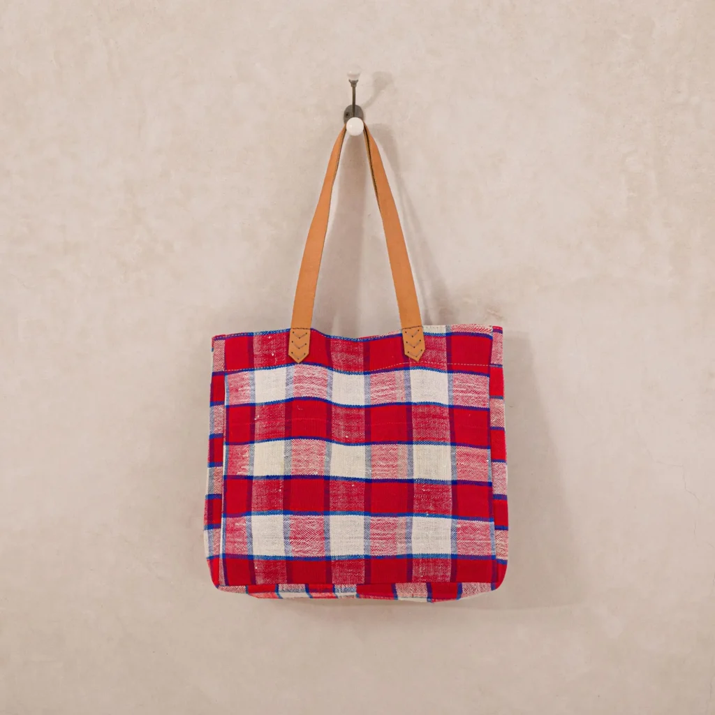 Red and white plaid tote bag with brown handles hanging on a hook against a beige wall.