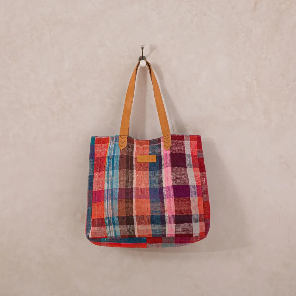 Colorful plaid tote bag with leather straps hanging on a wall hook against a light textured background.