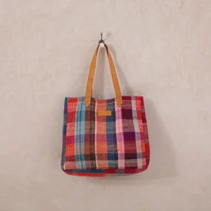 Colorful plaid tote bag with leather straps hanging on a wall hook against a light textured background.