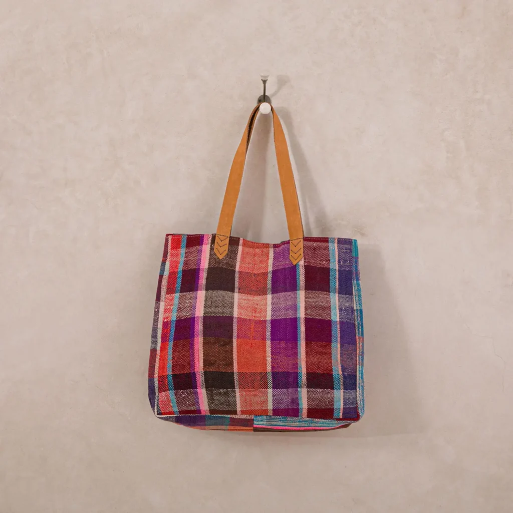 A colorful plaid tote bag with leather handles hangs on a wall hook.