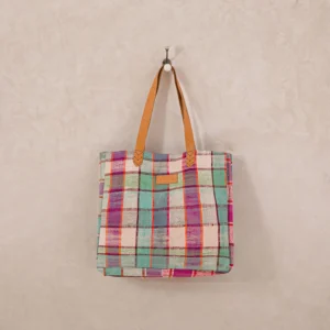 A colorful plaid tote bag with brown leather handles hangs on a wall hook against a textured beige background.