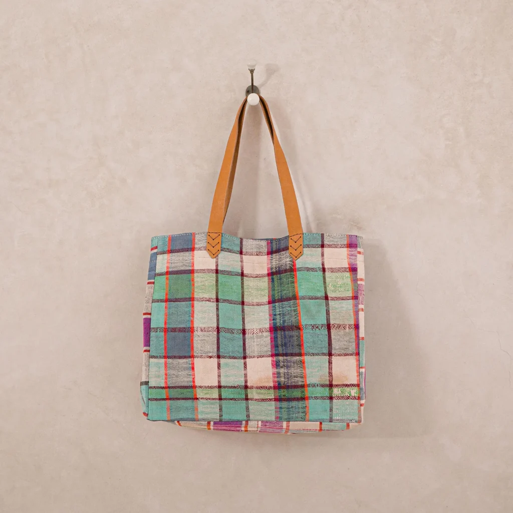 A multicolored plaid tote bag with brown handles hangs on a single wall hook against a beige background.