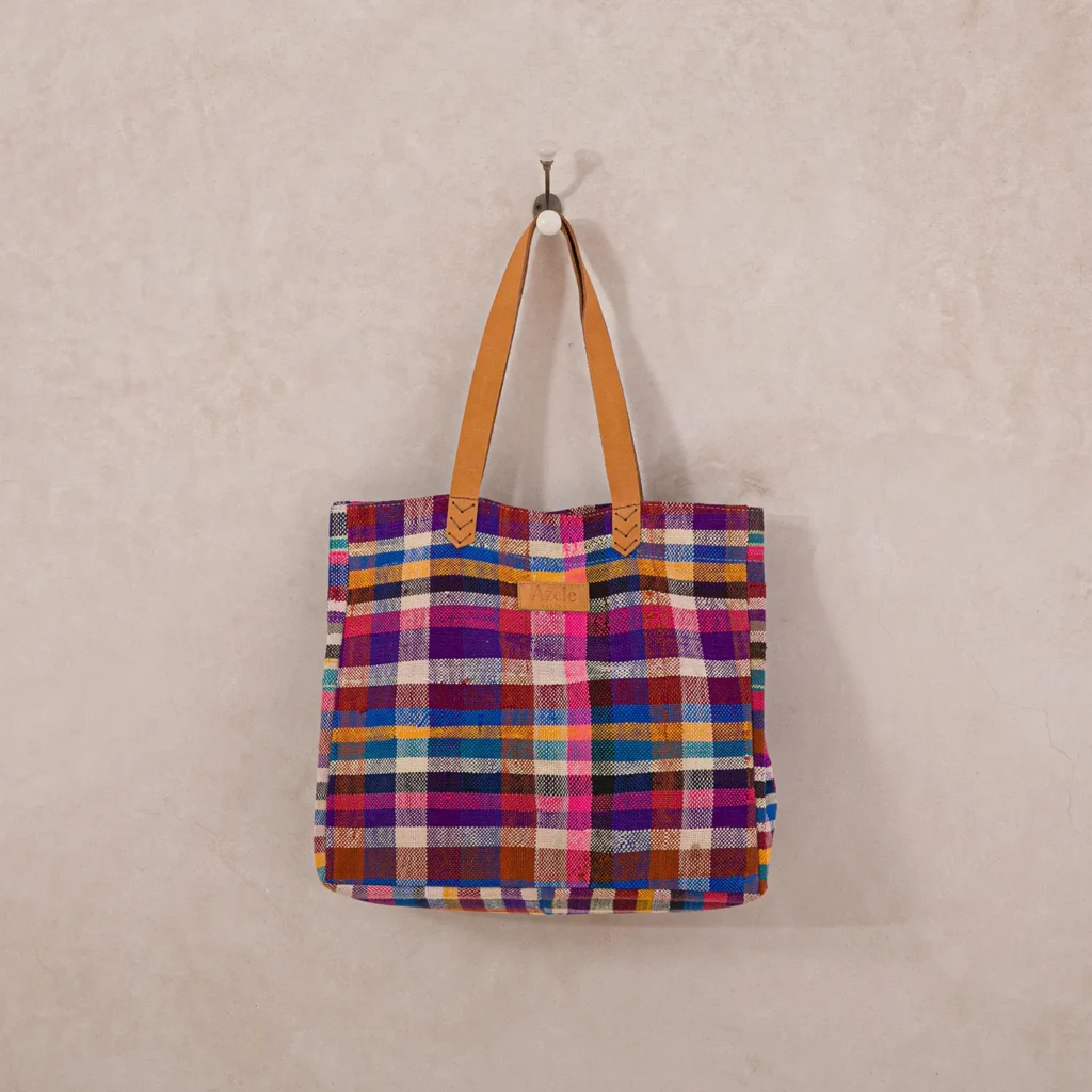 A multicolored plaid tote bag with brown leather handles hangs on a single metal hook against a light, textured wall.