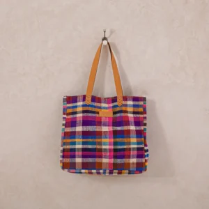 A multicolored plaid tote bag with brown leather handles hangs on a single metal hook against a light, textured wall.