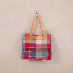 A colorful patchwork tote bag with tan handles hangs on a wall hook.