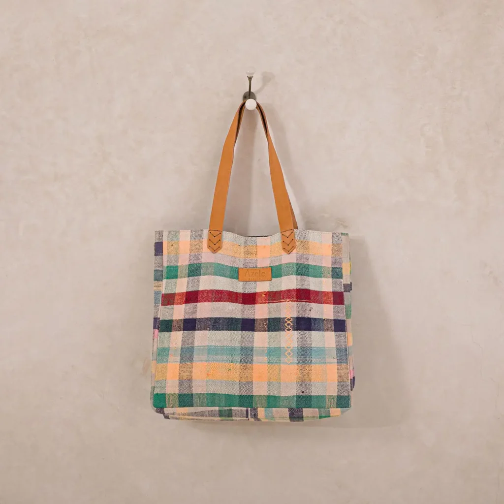 A multicolored plaid fabric tote bag with brown leather handles hangs on a wall hook.