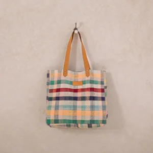 A multicolored plaid fabric tote bag with brown leather handles hangs on a wall hook.