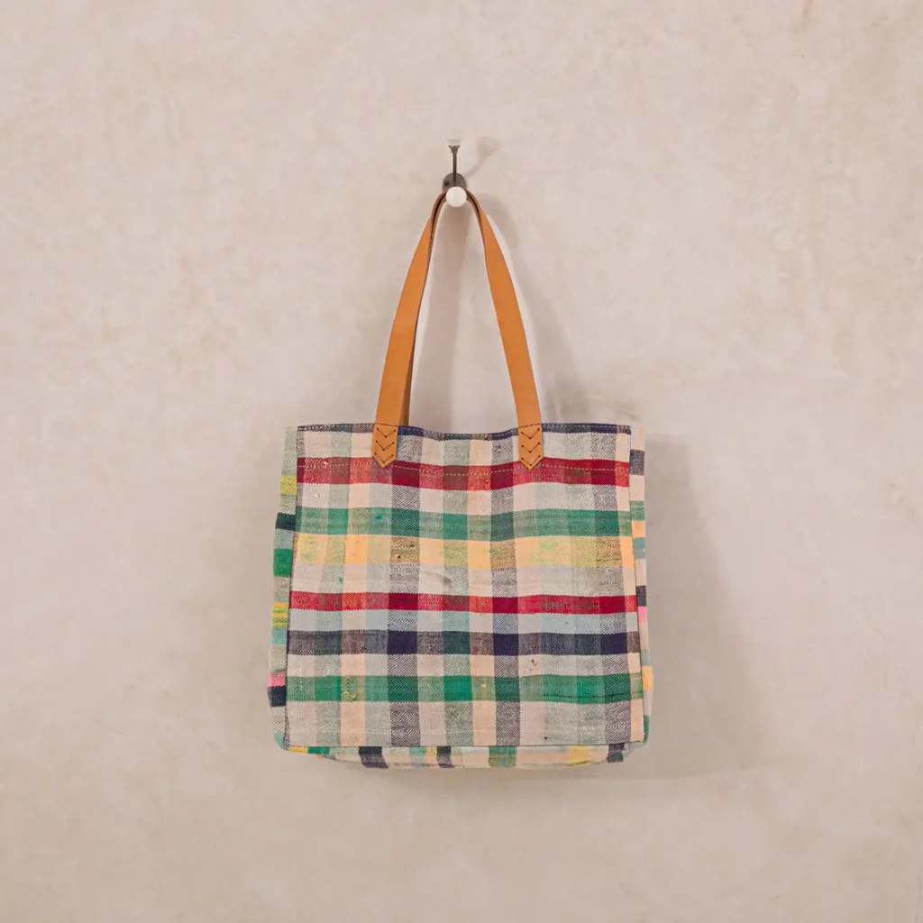 A plaid tote bag with brown leather handles hangs on a wall hook against a neutral background.