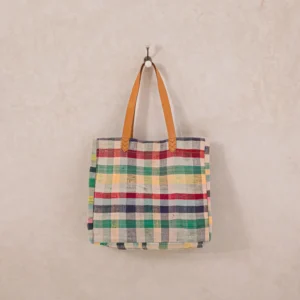 A plaid tote bag with brown leather handles hangs on a wall hook against a neutral background.