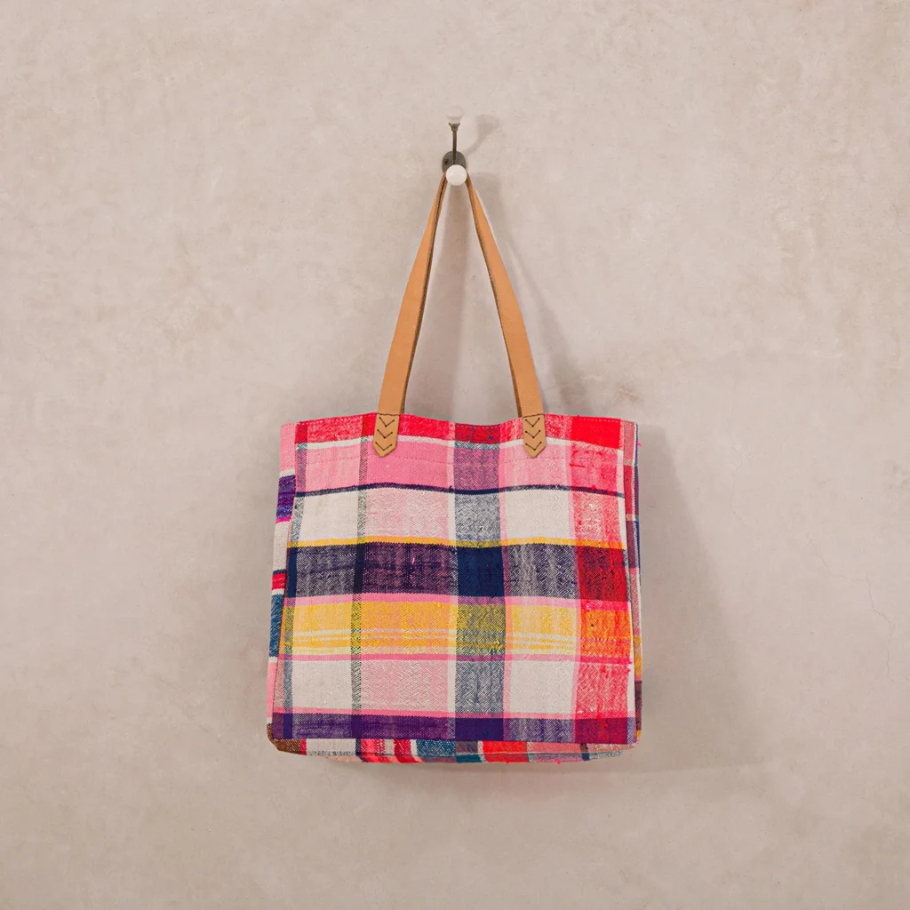 A colorful plaid tote bag with brown leather handles hangs on a wall hook.