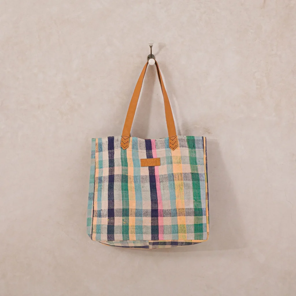A plaid patterned tote bag with brown leather handles hangs on a wall hook.