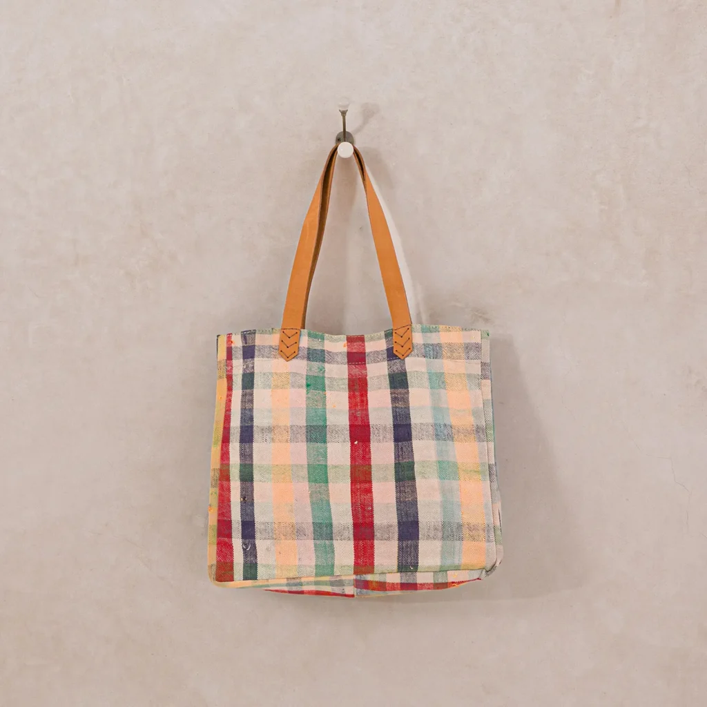 A plaid-patterned tote bag with tan leather handles hangs on a hook against a plain wall.