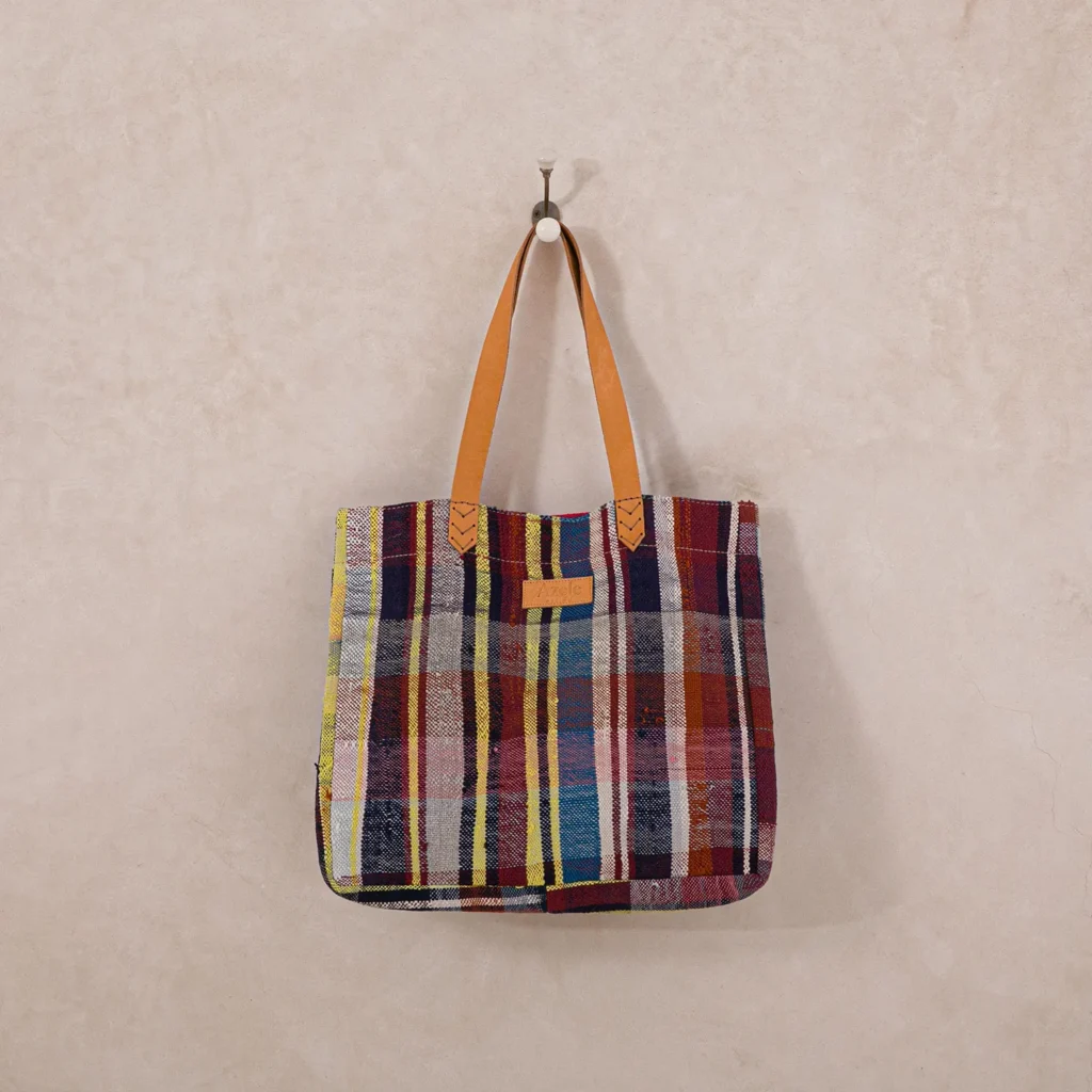 An upcycled multicolored plaid-patterned shopper bag with tan leather straps hangs on a single wall hook.