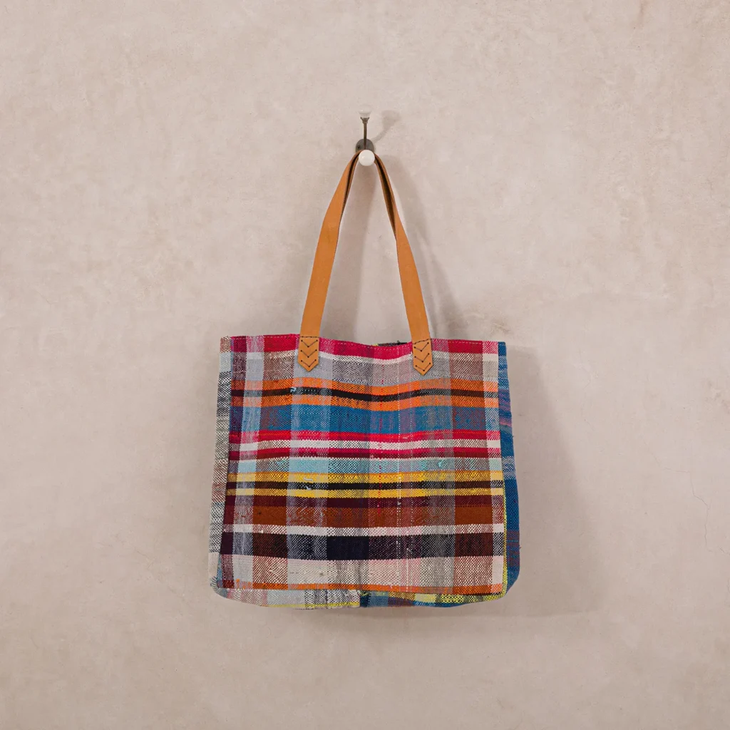 An upcycled shopper bag featuring a vibrant plaid pattern and brown leather handles hangs on a hook against a plain beige wall.