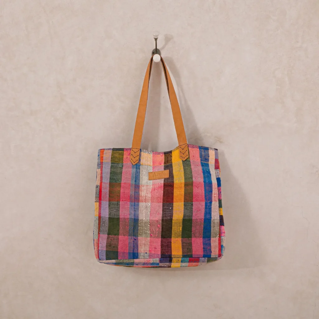 This vibrant upcycled shopper bag, featuring a colorful checkered design with brown leather straps, hangs elegantly on a wall hook.