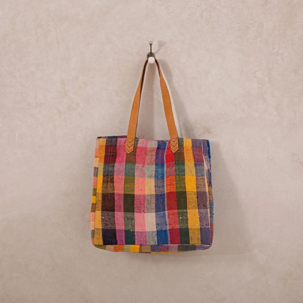 An upcycled shopper bag, multicolored and checkered, hangs from a wall hook, showcasing its two tan handles.