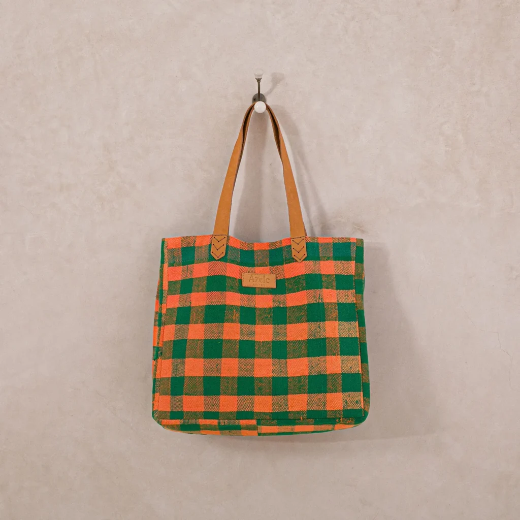 Upcycled plaid tote bag with a green and orange checkered pattern and brown leather handles, hanging on a hook.