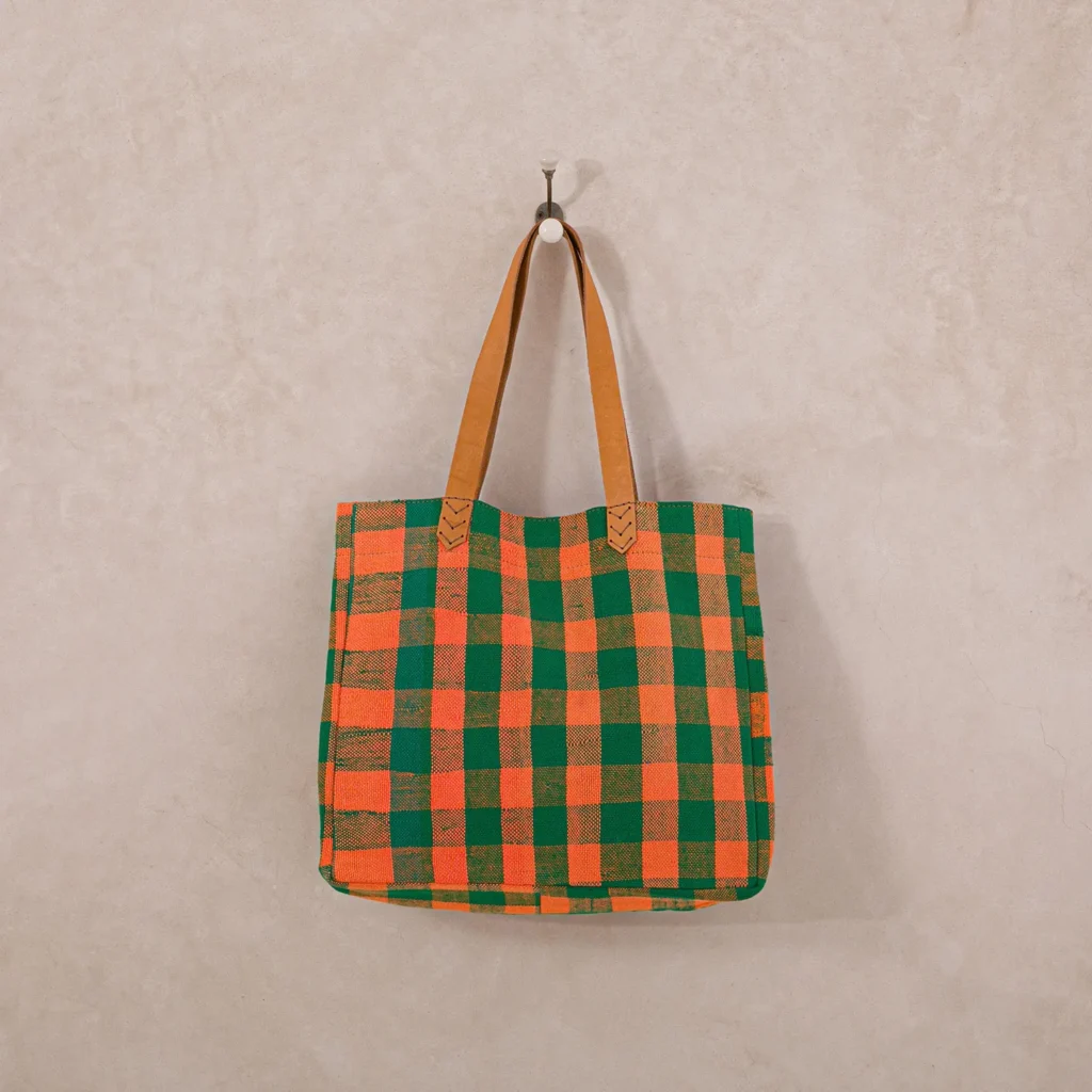An upcycled shopper bag, featuring vibrant orange and green checkered squares and brown leather handles, hangs elegantly on a textured beige wall.