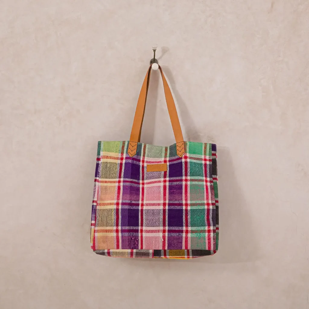 Plaid tote bag with multicolored squares and tan leather handles hanging on a wall hook against a neutral background.
