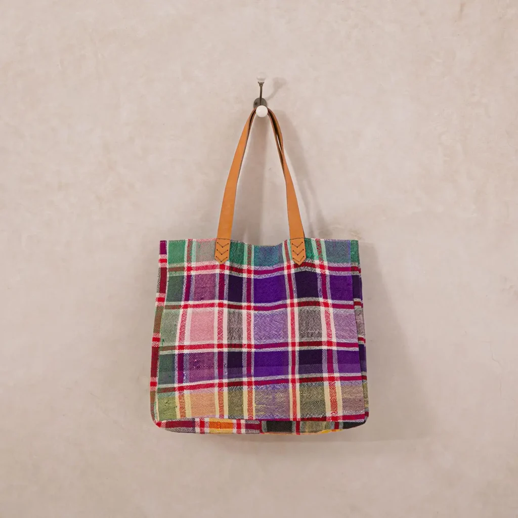 A multicolored plaid tote bag with tan leather handles hangs on a wall hook against a neutral background.