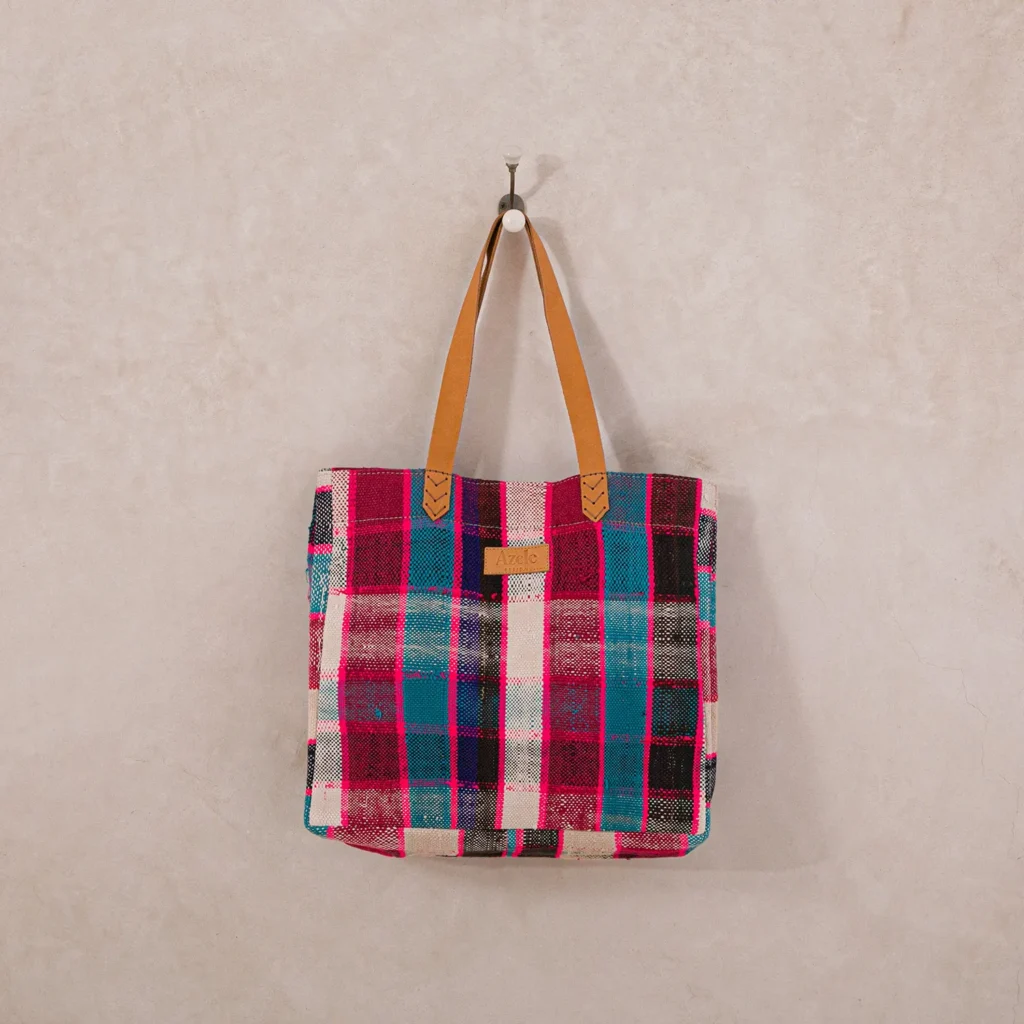 An upcycled shopper bag with pink, blue, and black checkered patterns hangs on a hook against a light-colored wall. This plaid tote features leather straps and a small label on the front, blending style with sustainability.