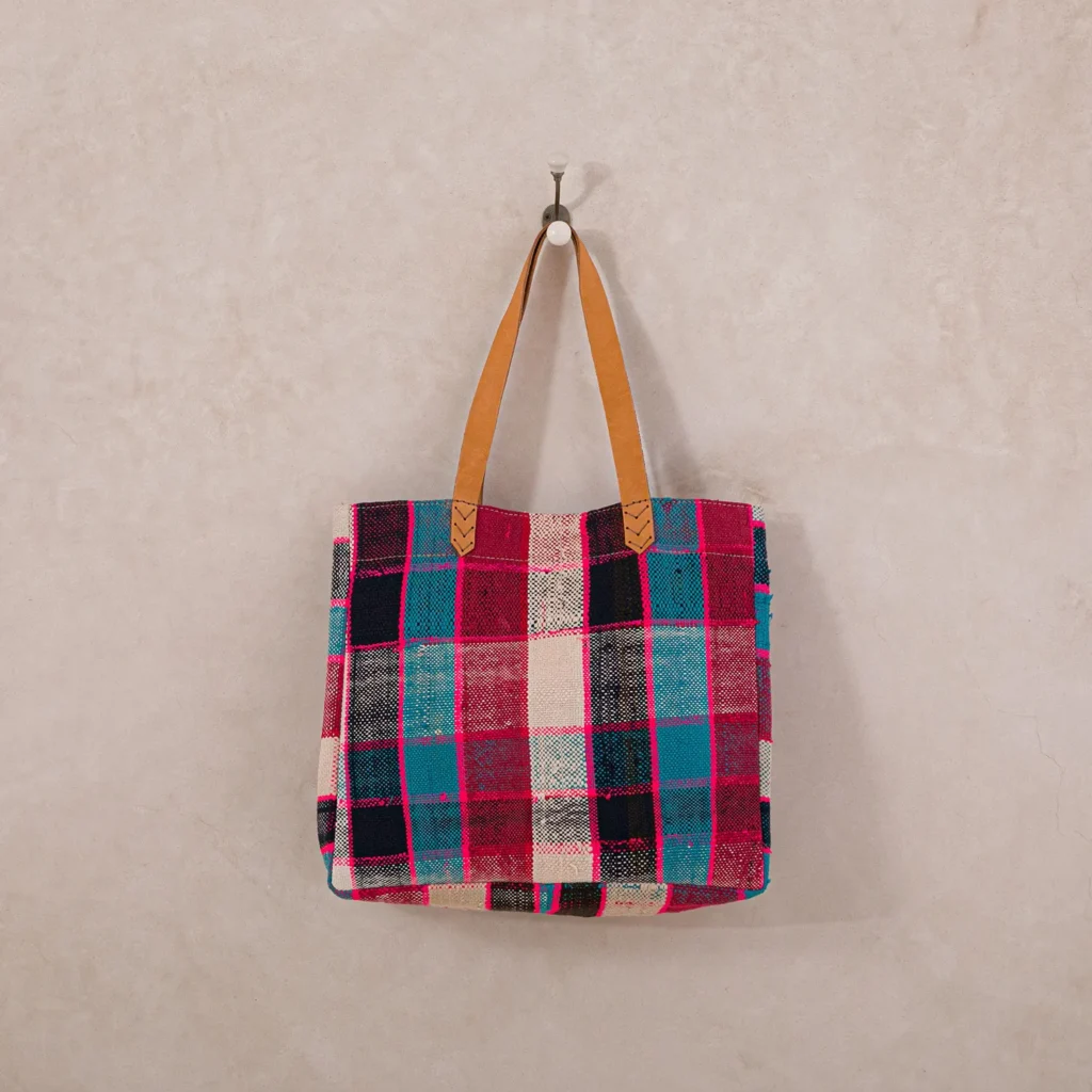 An upcycled shopper bag with a red, blue, and white checked plaid pattern hangs on a wall hook. The tote is adorned with elegant tan leather handles.