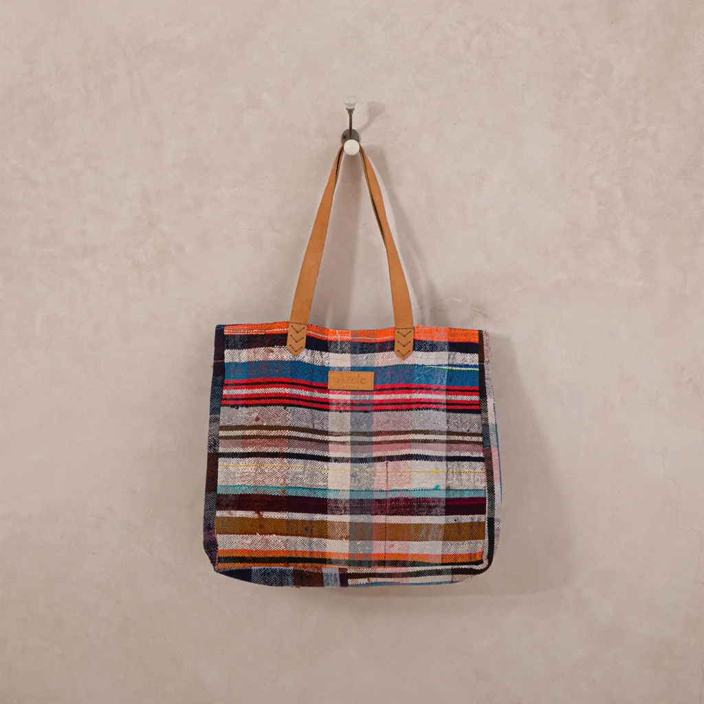 Multicolored striped fabric tote bag with leather handles hangs on a hook against a light beige wall.