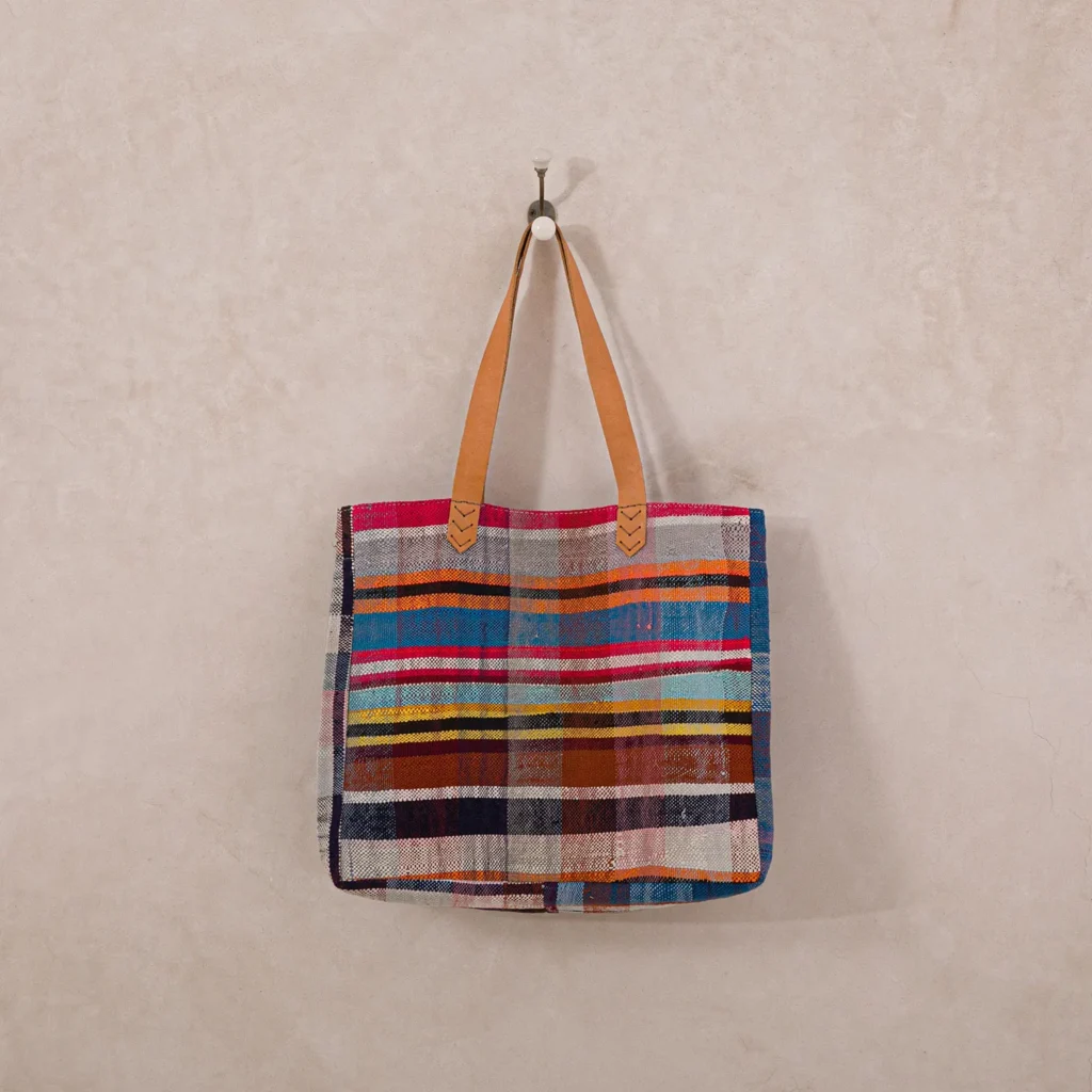 Colorful striped fabric tote bag with tan leather handles hanging on a wall hook.