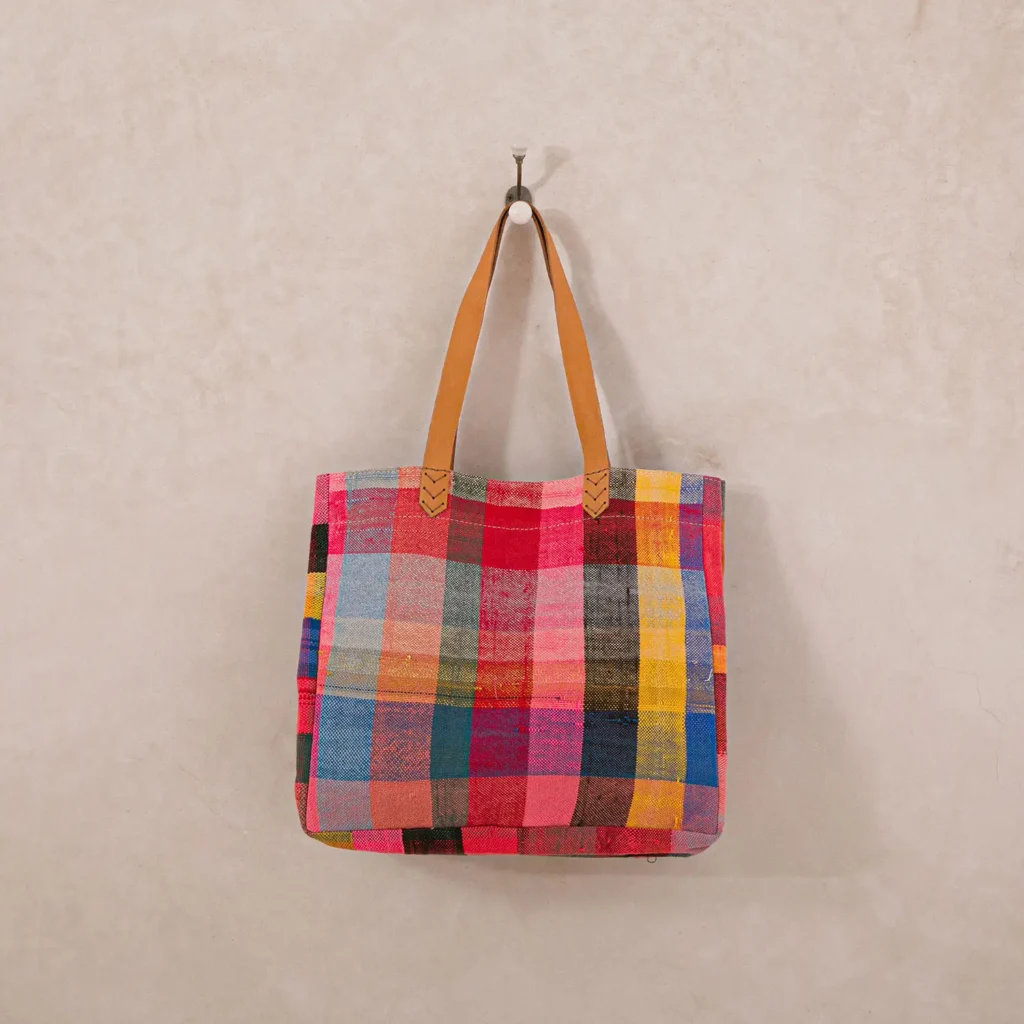 A multicolored checkered tote bag with leather handles hangs on a hook against a plain wall.