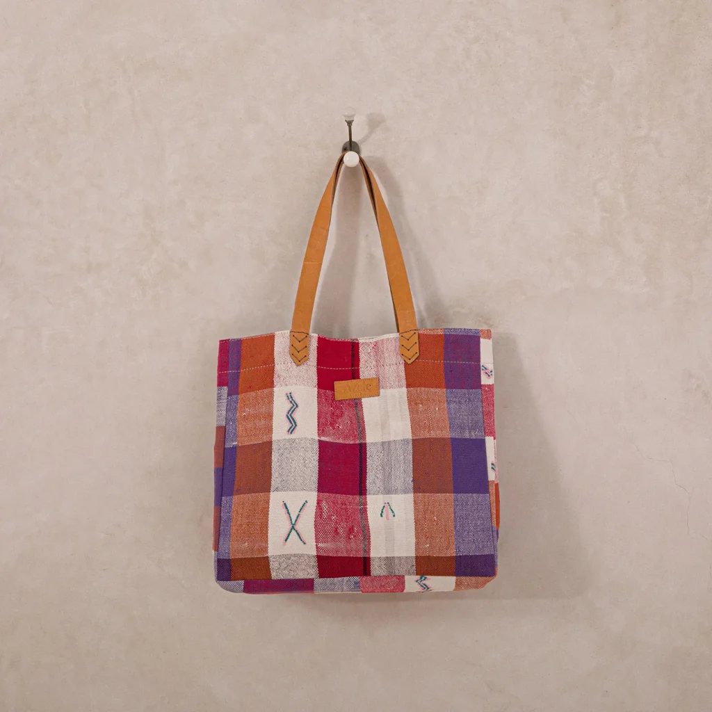 Patchwork tote bag with brown leather handles hangs against a light textured wall. The bag features a checkered design with various colors and abstract patterns.
