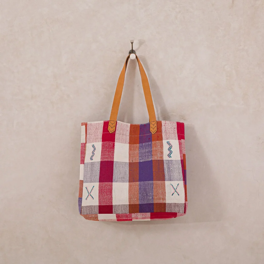 A woven tote bag with a red, white, and blue plaid pattern, featuring brown leather handles, hangs on a wall hook.