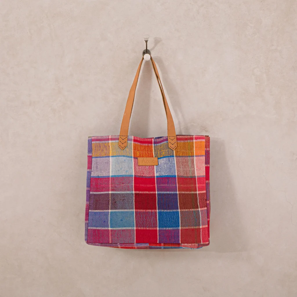A colorful patchwork tote bag with tan leather handles hangs on a simple wall hook. The bag features a variety of vibrant colored squares.
