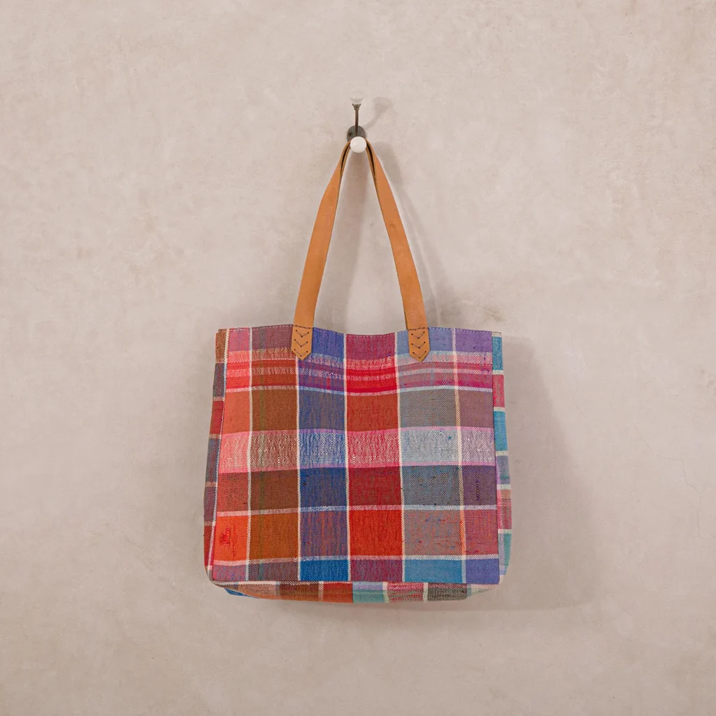 A colorful patchwork tote bag with leather handles hangs on a single wall hook against a beige wall.