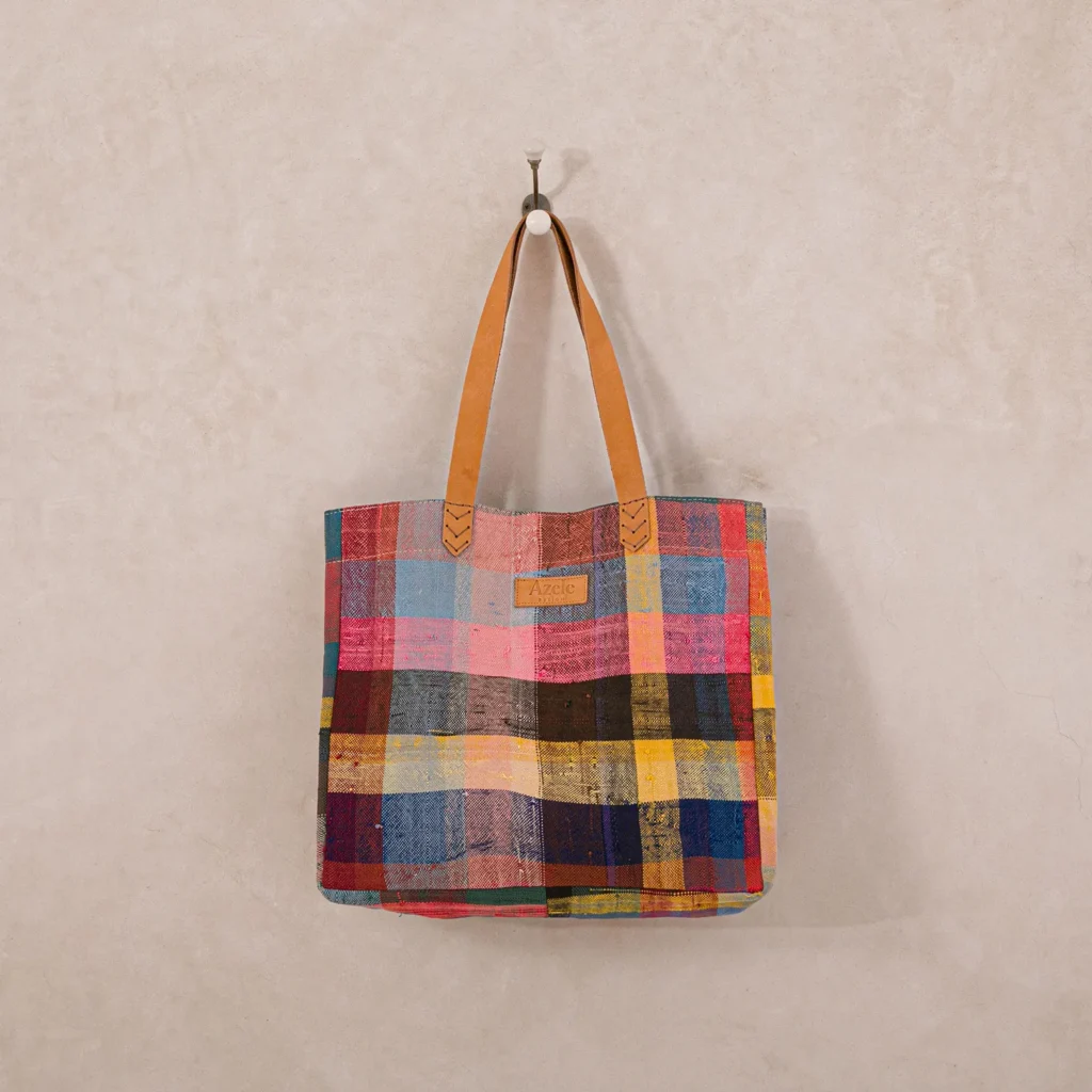 Colorful plaid tote bag with tan leather straps hanging on a single hook against a plain wall.