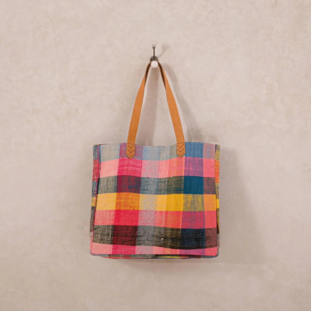 A multicolored plaid tote bag with brown leather handles hangs on a wall hook.