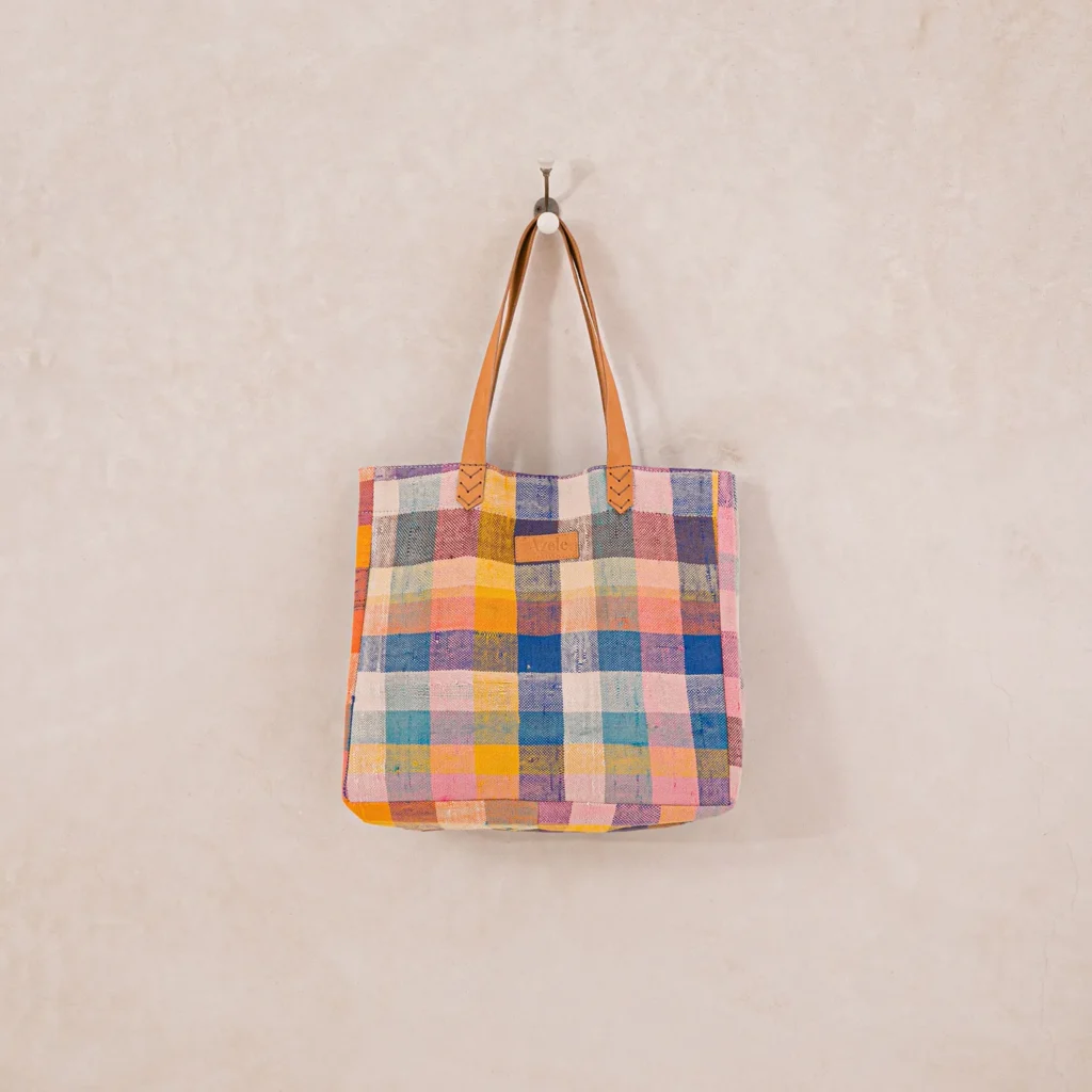 A multicolored, checkered tote bag with leather handles hangs on a wall hook against a light beige background.