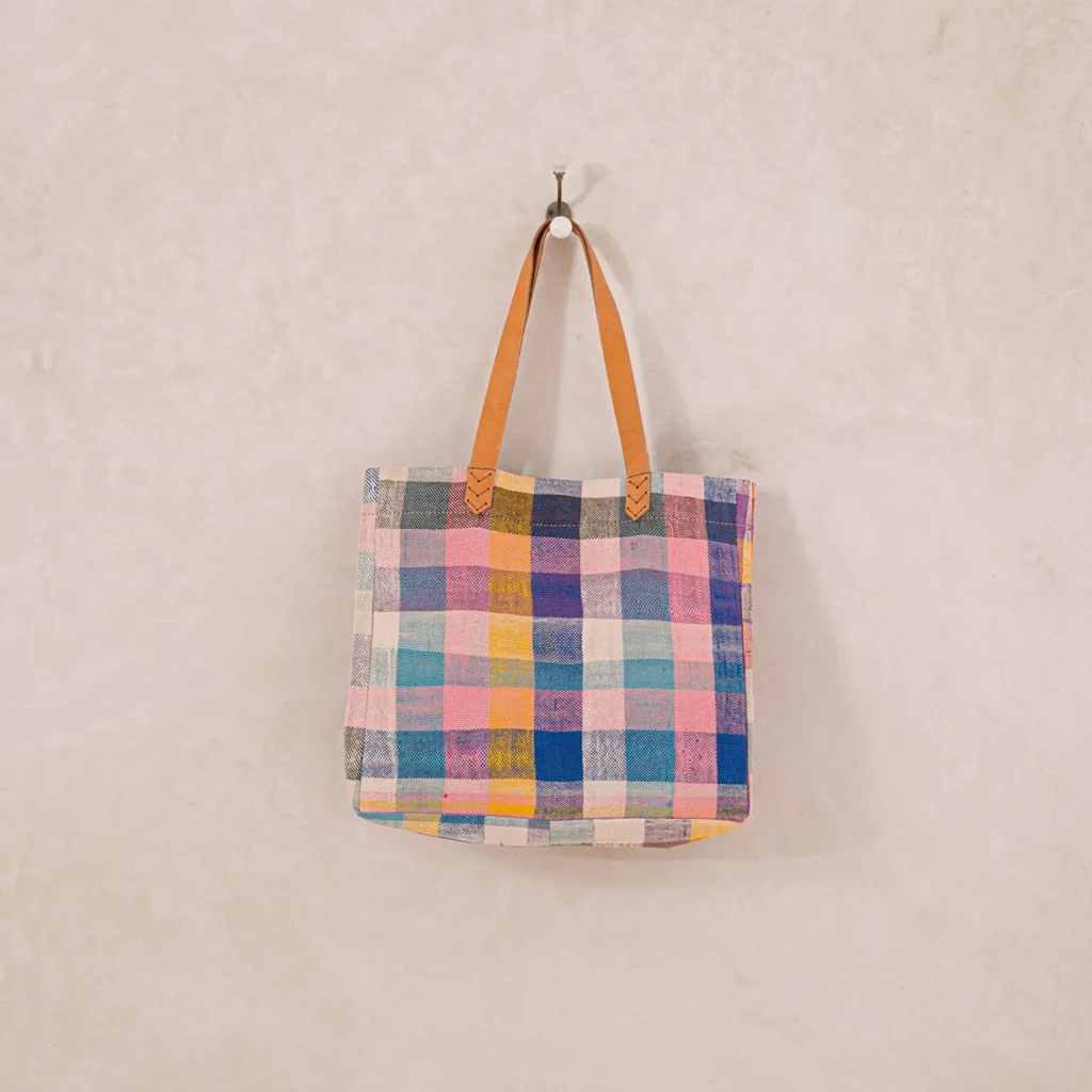 A multicolored plaid tote bag with brown leather handles hangs on a wall hook.