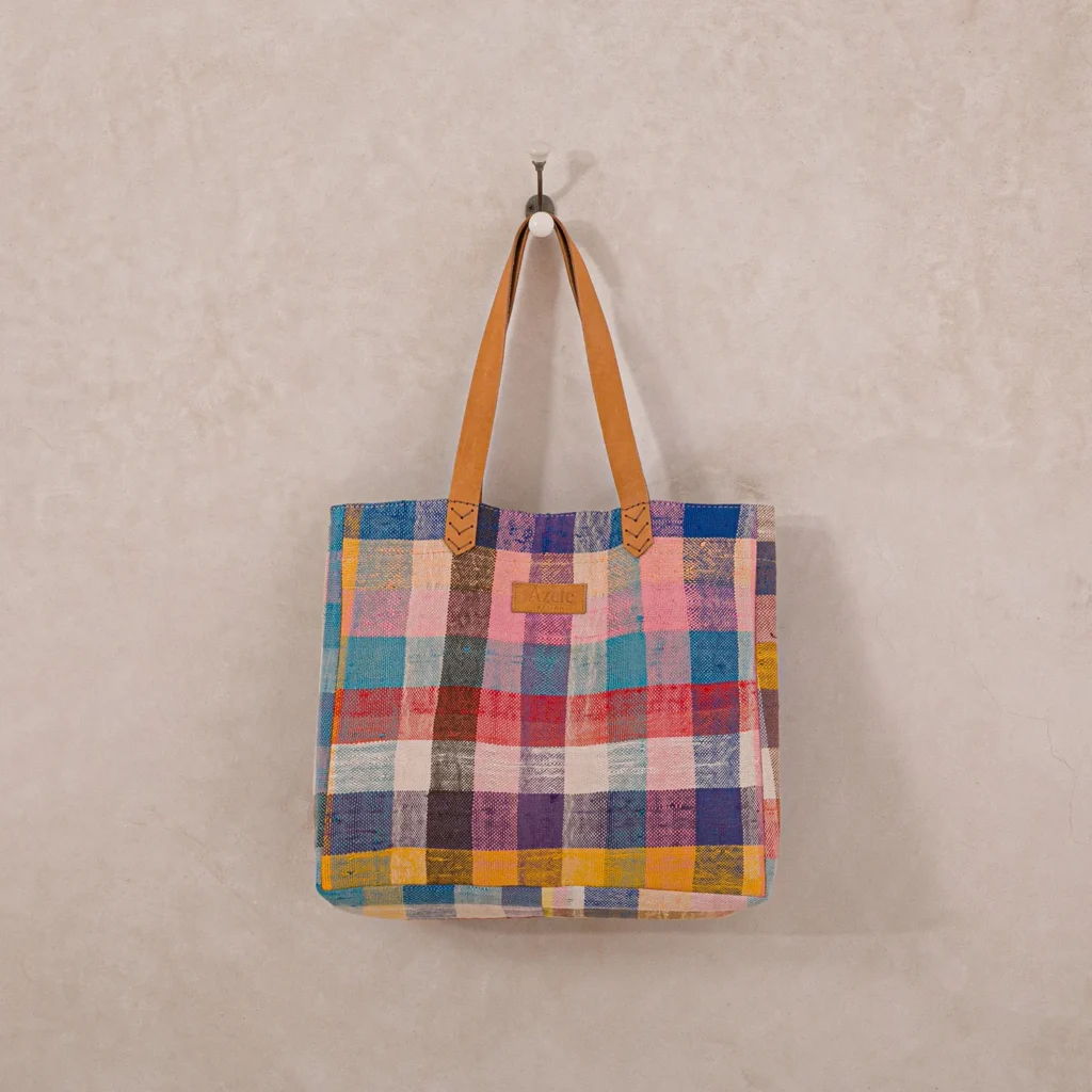 A multicolored plaid tote bag with brown leather straps hangs on a wall hook against a beige background.