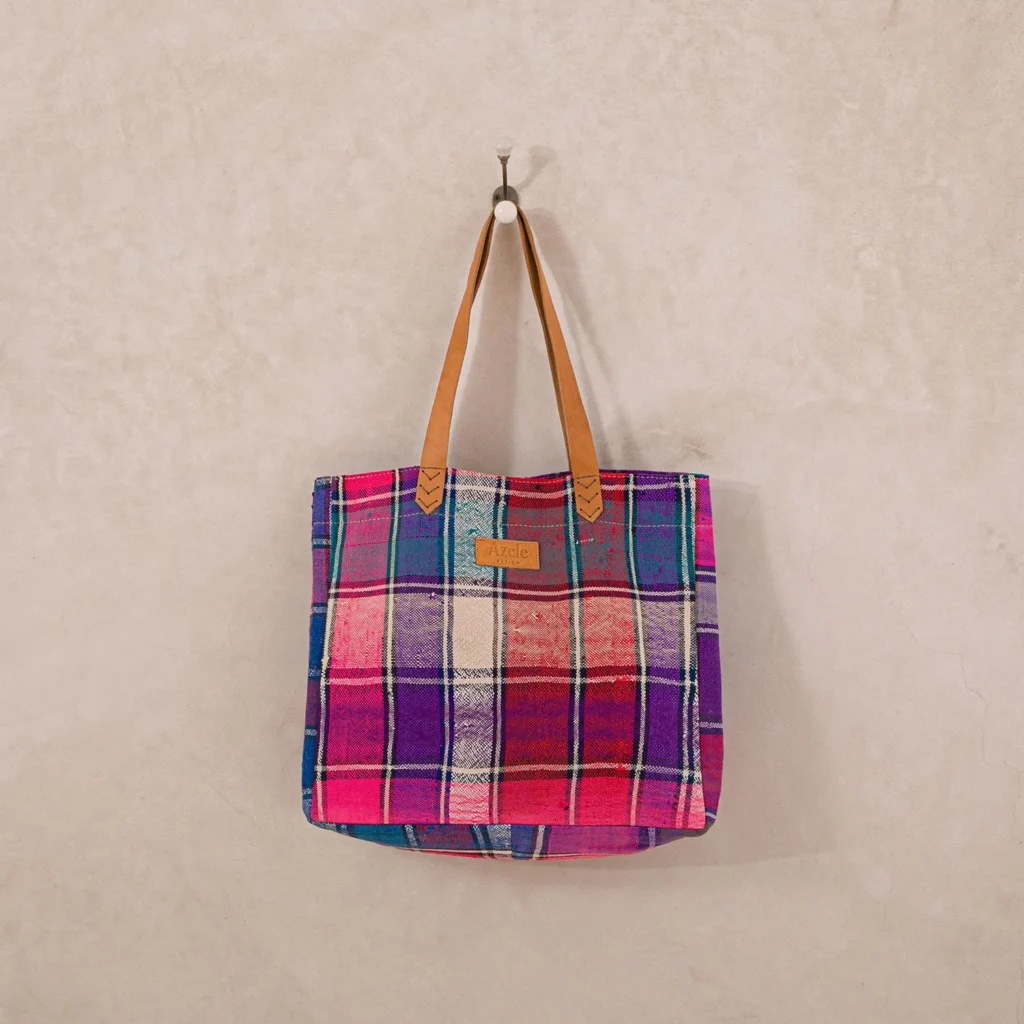 A plaid tote bag with leather handles is hanging on a wall hook. It features pink, purple, and red checkered patterns.