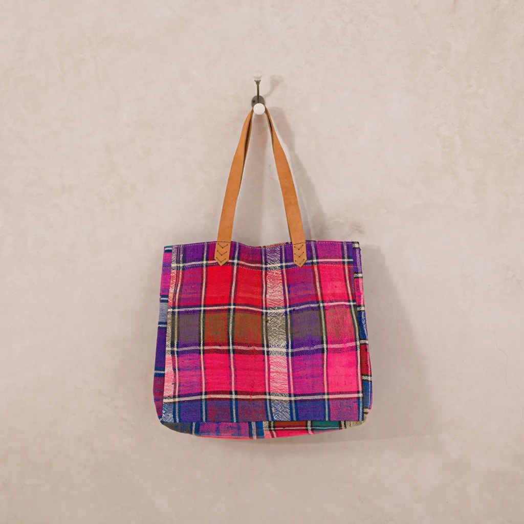 A colorful plaid tote bag with brown leather handles hangs on a simple wall hook against a beige wall.