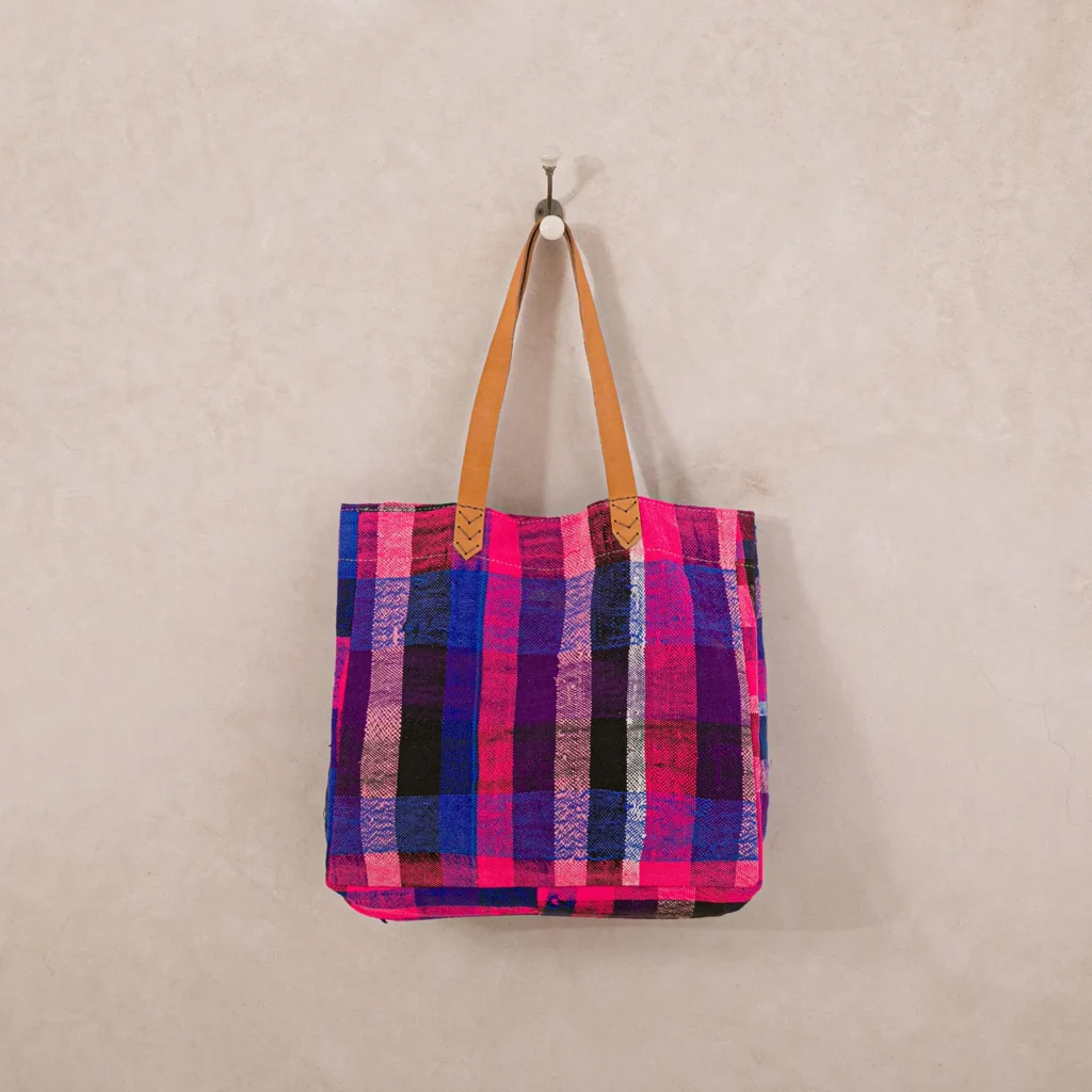 A colorful plaid tote bag with tan leather handles hangs on a wall hook.