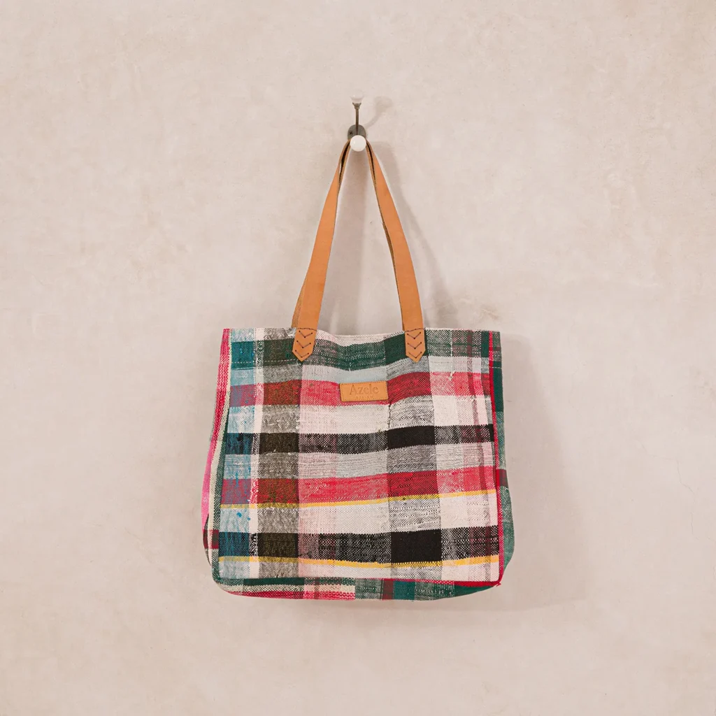 A multicolored plaid tote bag with tan leather handles hangs on a metal hook against a textured beige wall.
