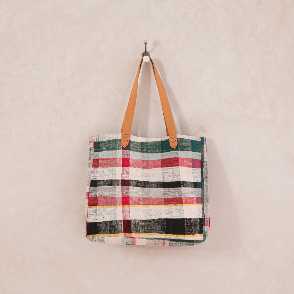 A multicolored plaid tote bag with tan handles hangs on a wall hook.