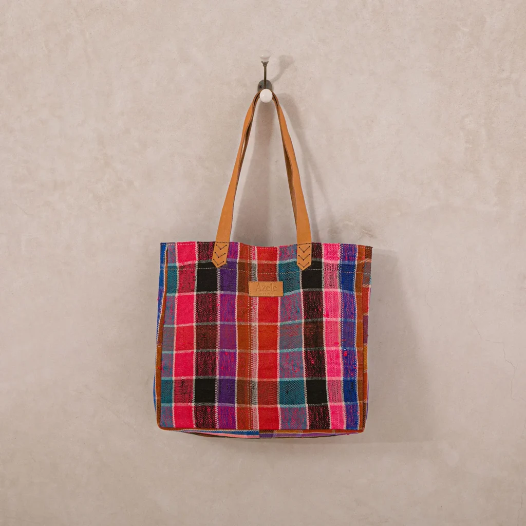 A multicolored plaid tote bag with leather straps hangs on a wall hook.
