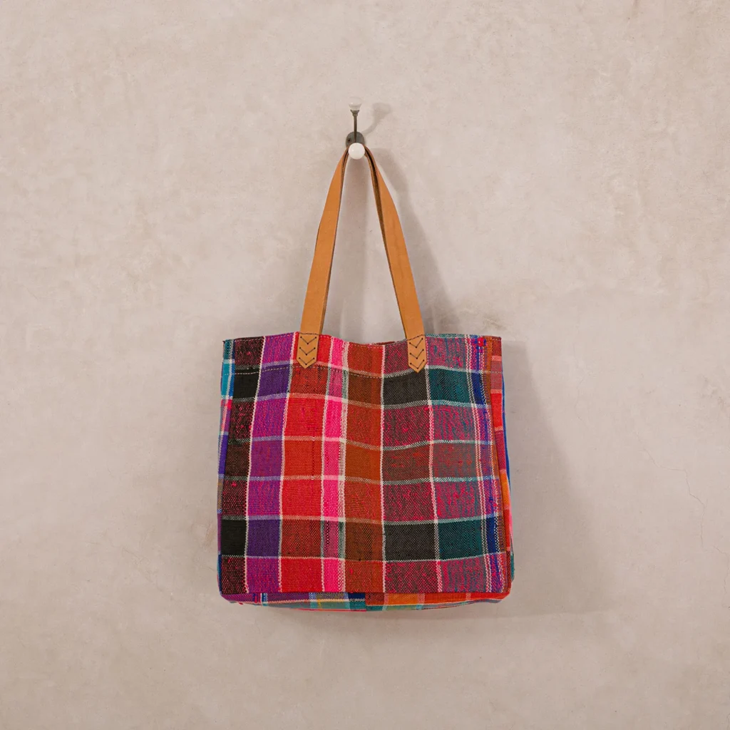 A colorful plaid tote bag with leather handles hangs on a wall hook.
