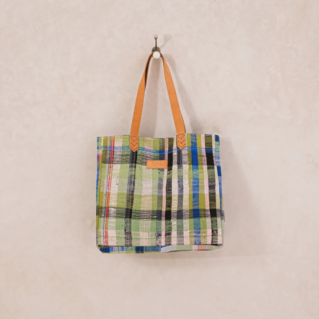A plaid tote bag with green, blue, and red patterns hangs on a wall hook. It has brown leather handles and a small leather patch on the side.