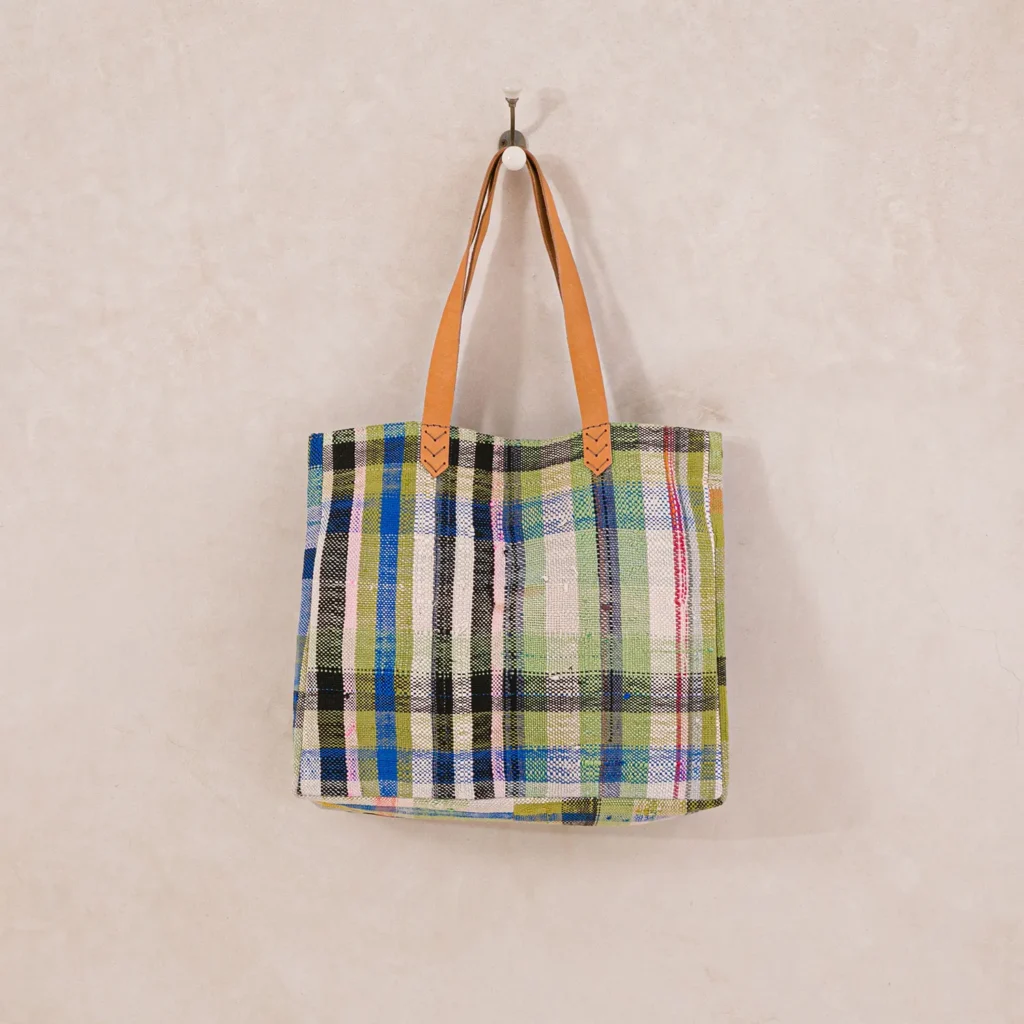 A multicolored plaid tote bag with tan leather handles hangs on a wall hook.